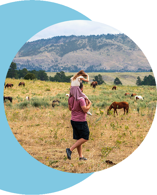 A man walking through an open field with his daughter on his shoulders.