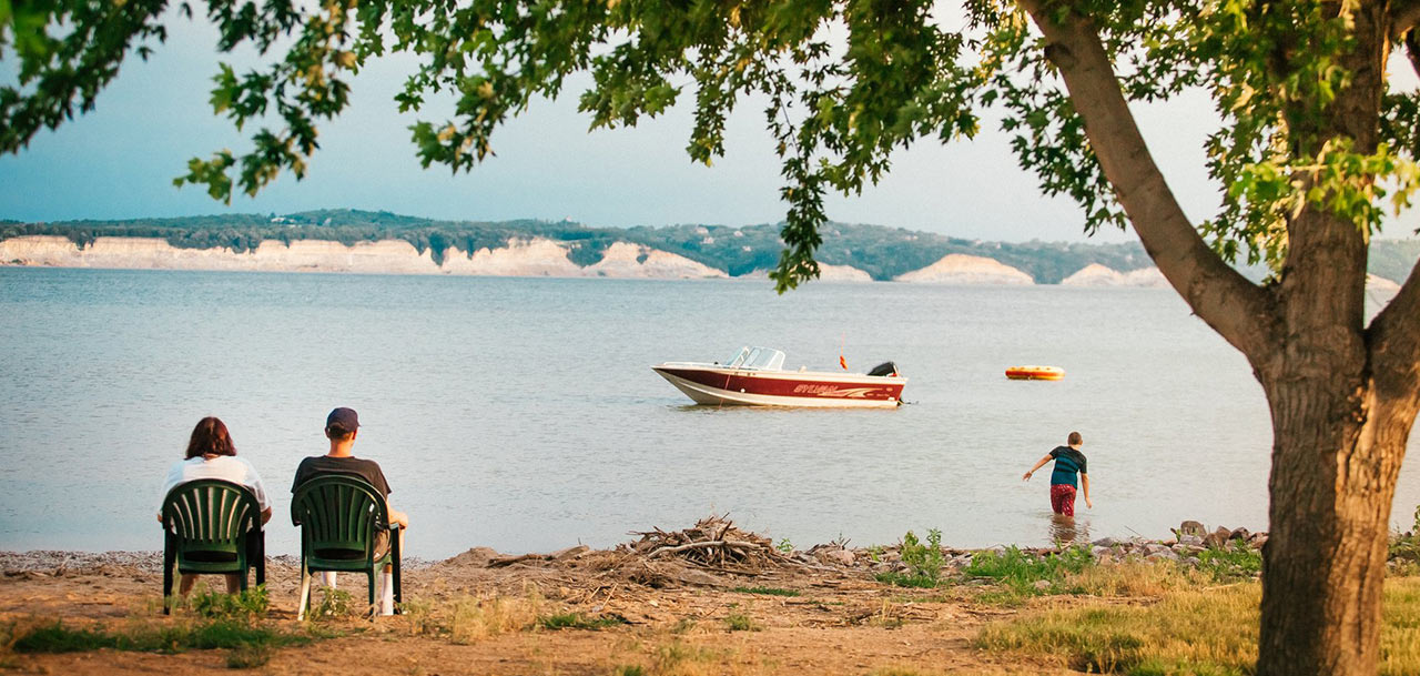 A mother and father sit by a lake while their son wades in the water.