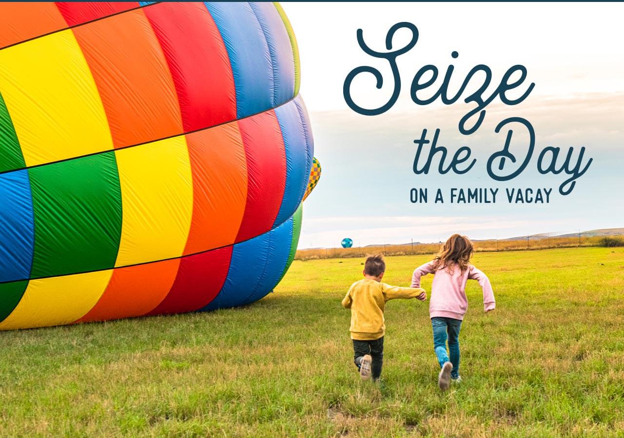 Two kids running in a field along side a colorful hot air balloon that is on the ground.