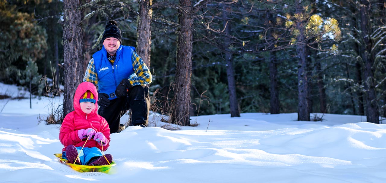 A father watching his young daughter on a child's sled.