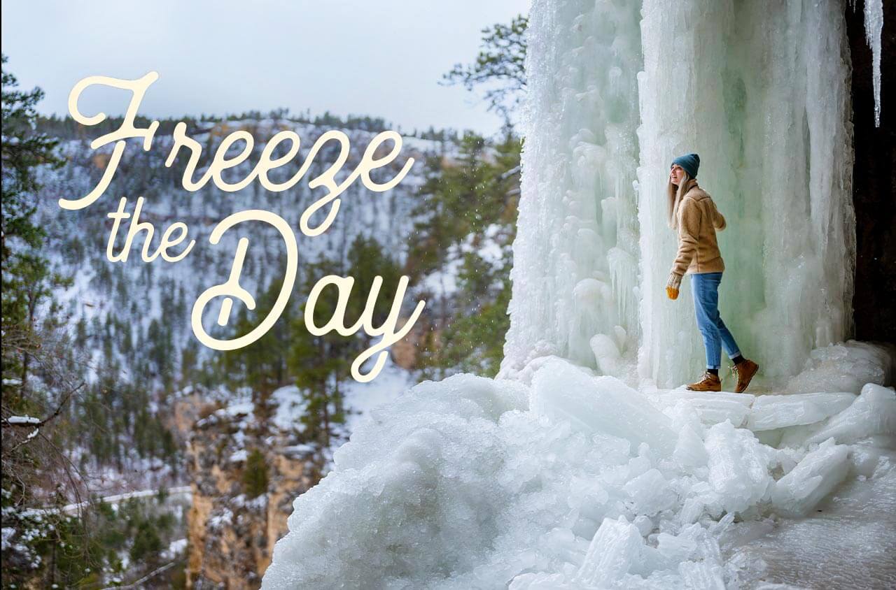 A girl standing under a frozen waterfall overlooking a snow covered forest.