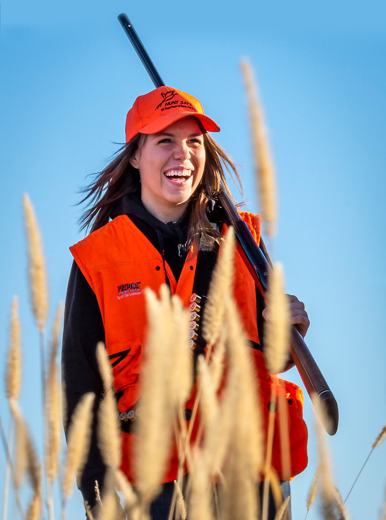 A woman smiling with a gun resting on her shoulder as she's out walking through a wheat field while hunting.